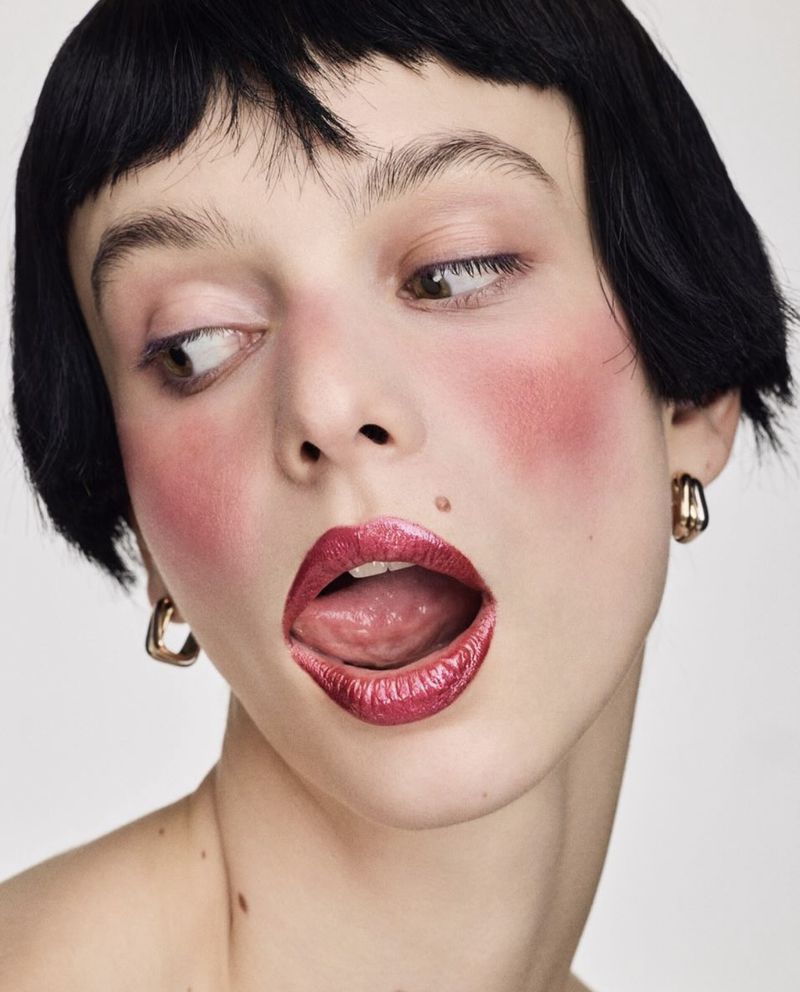 Close-up portrait of a young adult woman with black hair, pink blush, and bright red lipstick, sticking her tongue out against a white backdrop.