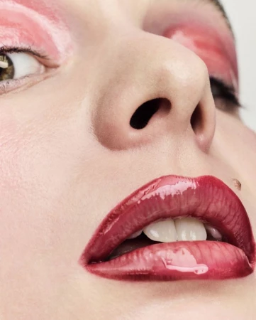 Extreme close-up portrait of a model with glossy pink eyeshadow and dark red glossy lips against a neutral background.