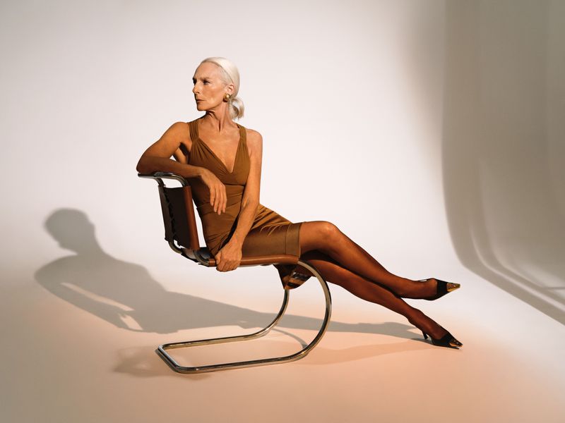 Elderly female model in brown dress seated on a modern chrome chair against a seamless white studio backdrop