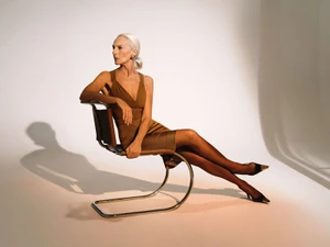 Elderly female model in brown dress seated on a modern chrome chair against a seamless white studio backdrop
