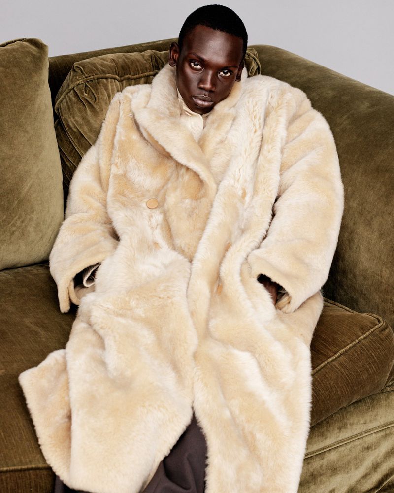 Person with dark skin and an off-white coat lying on a green couch, looking straight ahead into the camera in a portrait-style shot.