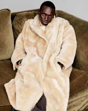 Person with dark skin and an off-white coat lying on a green couch, looking straight ahead into the camera in a portrait-style shot.