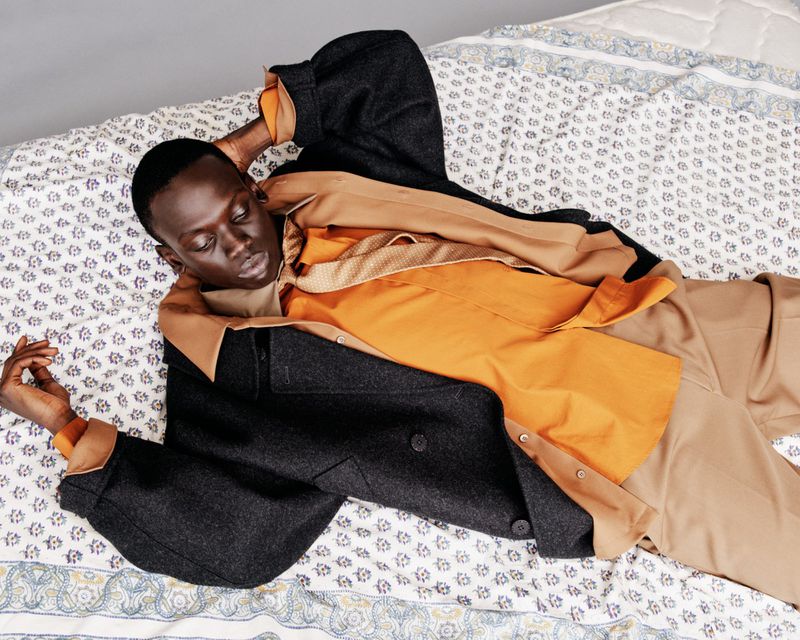 Dark-skinned male model in a casual pose on a bed with patterned sheets, wearing a button-down shirt and jacket in a bright, eye-level shot.