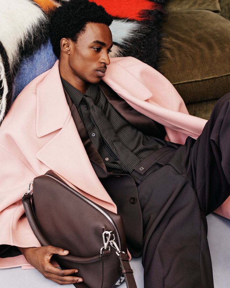 Man in pink coat sitting with a brown leather bag in a fashion editorial, styled with a dark shirt and black pants, studio lit.