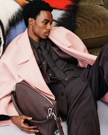 Man in pink coat sitting with a brown leather bag in a fashion editorial, styled with a dark shirt and black pants, studio lit.