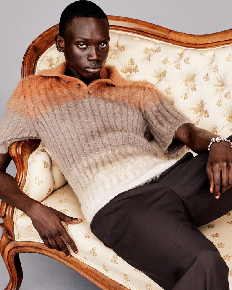 Dark-skinned man in brown sweater reclines on vintage couch. Fashion portrait with relaxed pose in a warm-toned indoor setting.