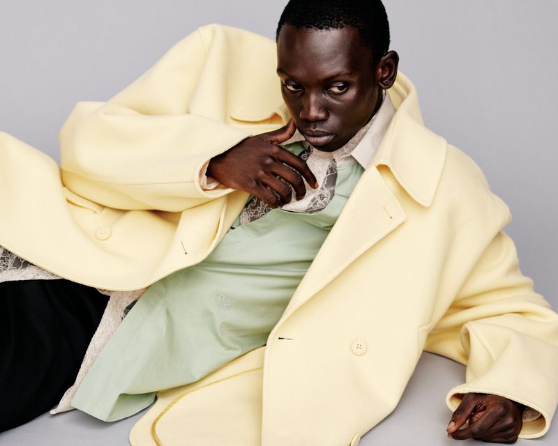 Dark-skinned man in a pale yellow coat poses in a studio setting, looking intently at the camera with a neutral expression.