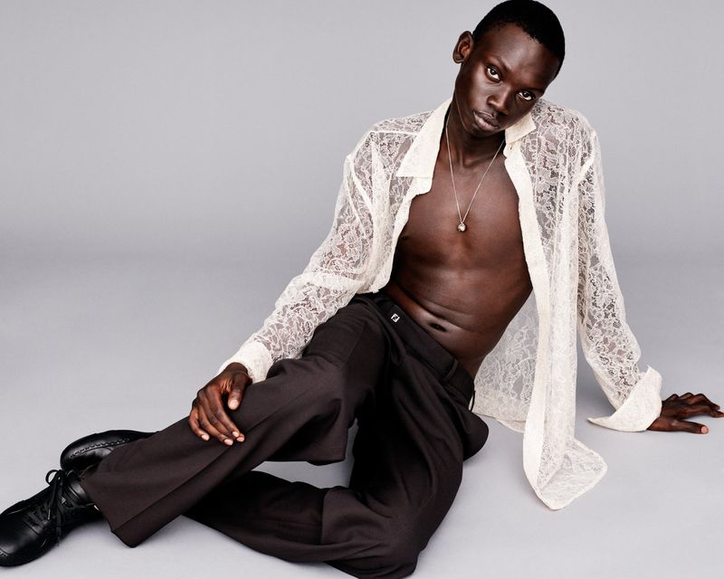 An adult male fashion model with dark skin sits on a grey backdrop wearing black pants and a white lacy button-up shirt with an open collar.