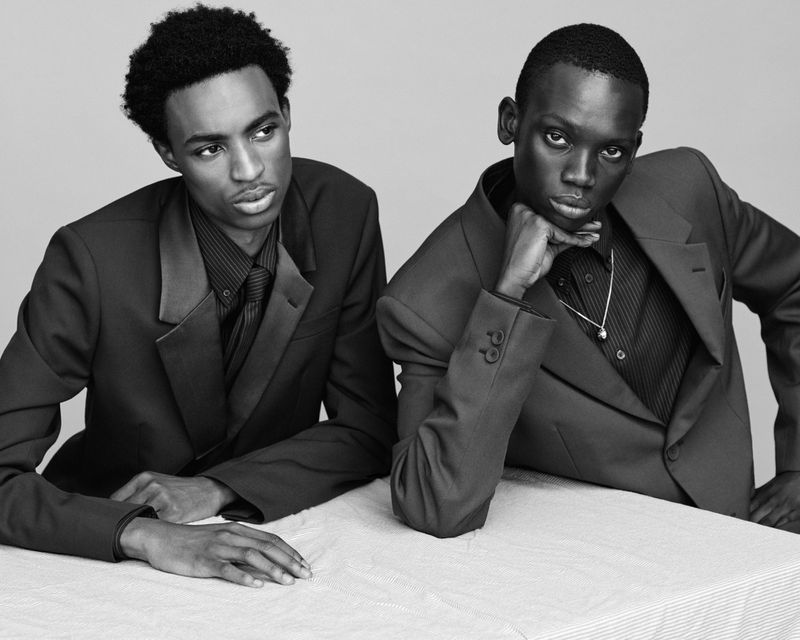 Two Black men pose indoors wearing suits and button-down shirts, leaning on a table in a studio setting with a neutral gray background.