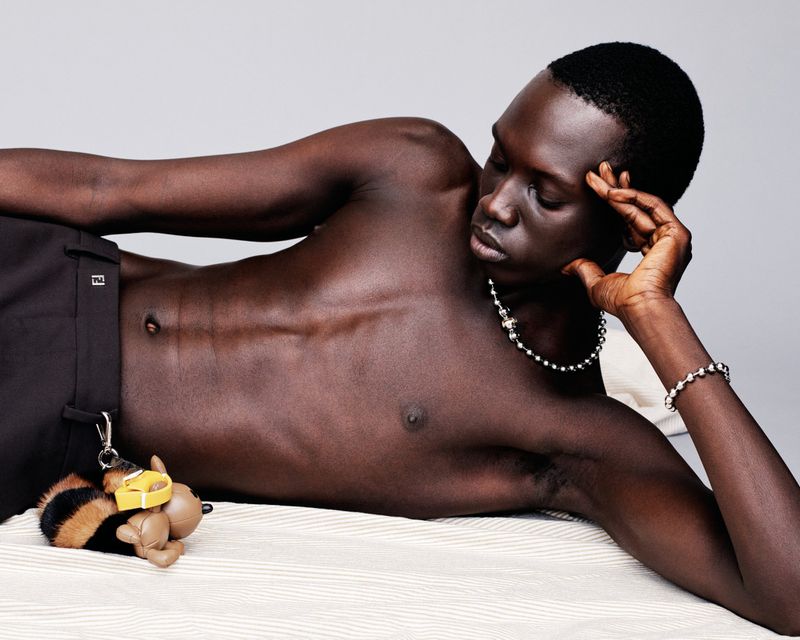 Man lying on a bed wearing jewelry and black pants, accessorized with a bee-shaped keychain, in a photo studio setting.