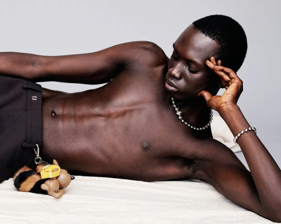 Man lying on a bed wearing jewelry and black pants, accessorized with a bee-shaped keychain, in a photo studio setting.