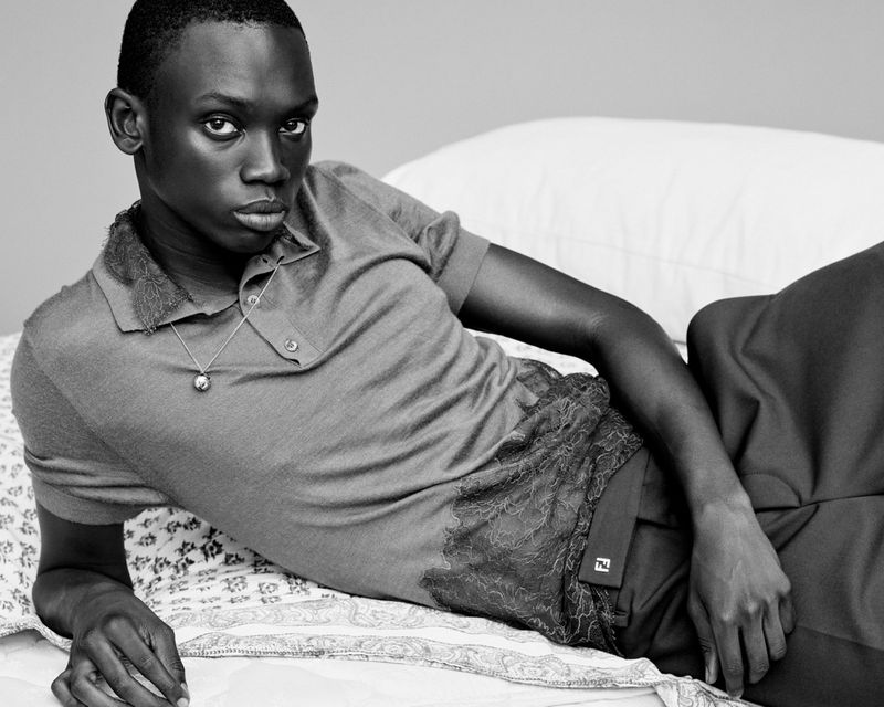 Portrait of a Black male model in a polo shirt and dark trousers lying on a floral patterned bed with a white pillow against a grey wall.