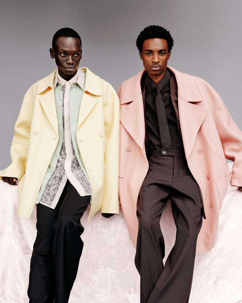 Two African-American men pose in pastel coats and dark trousers against a grey backdrop, blending fashion and contemporary style.