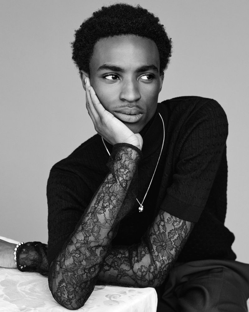 Portrait of a black male with dark hair in a black turtleneck and lace sleeves, resting his face on his hand in a studio setting.