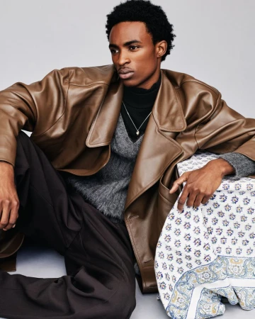 Young Black model wearing a brown leather coat and dark trousers in a studio setting by photographer Clay Stephen Gardner.
