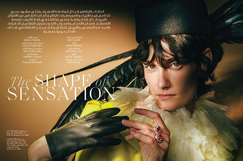 Close-up portrait of a model wearing a black feathered hat, ruffled yellow collar, black gloves and a ruby ring, indoors.