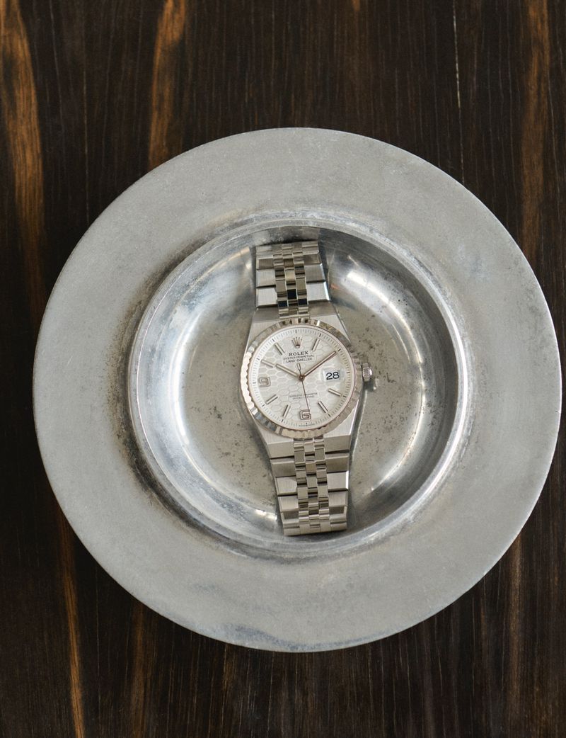 Silver wristwatch with a jubilee bracelet on a silver plate, positioned on a dark brown wooden table, overhead shot.