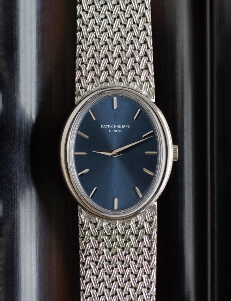 Close-up studio shot of a Patek Philippe watch with a blue face and silver braided wristband, resting on a dark textured background.