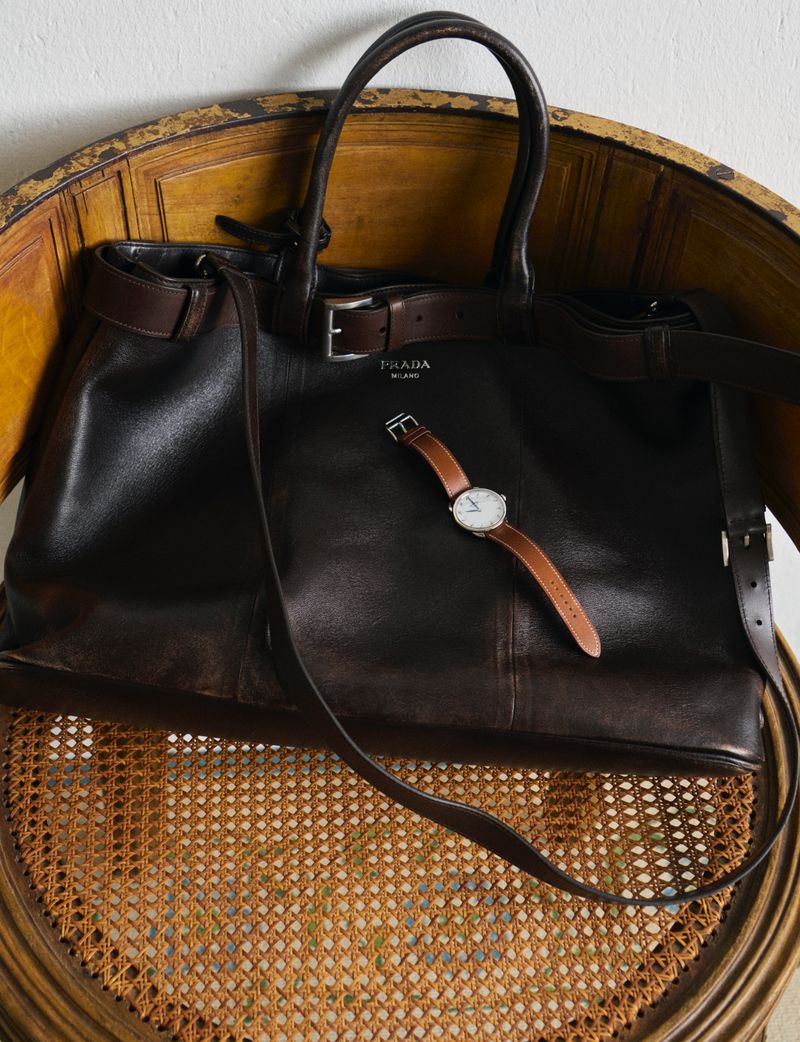 Dark brown leather Prada handbag and brown leather watch are arranged on a vintage cane chair against a white wall.