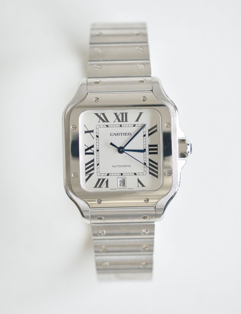 Elevated studio shot of a silver Cartier Santos wristwatch with a square face, Roman numerals, and blue hands on a white background.