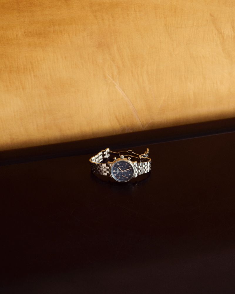 Close-up studio shot of a stainless steel chronograph watch with a navy dial, displayed on a reflective surface with a wooden backdrop.