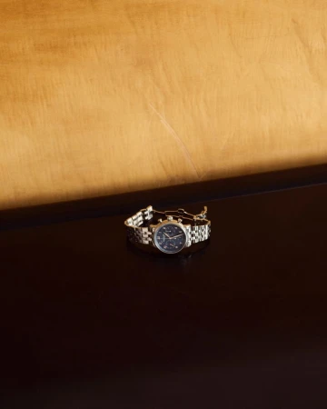 Close-up studio shot of a stainless steel chronograph watch with a navy dial, displayed on a reflective surface with a wooden backdrop.
