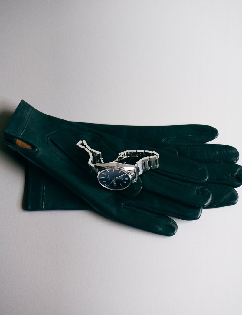 Luxury wrist watch with a silver wristband laying on a pair of dark green leather gloves against a white background in a studio setting.
