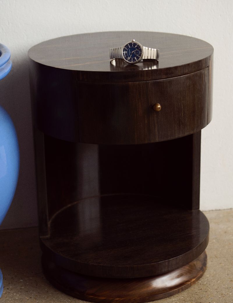 Wristwatch sits atop a dark wood bedside table with a drawer; a blue vase is partially visible to the left in an indoor setting.