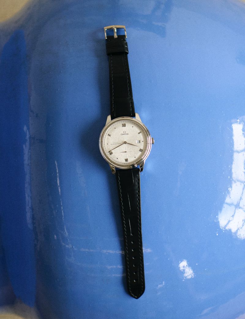 Elevated shot of a silver Omega wristwatch with a black leather strap, positioned on a glossy blue surface. Focus on timekeeping.