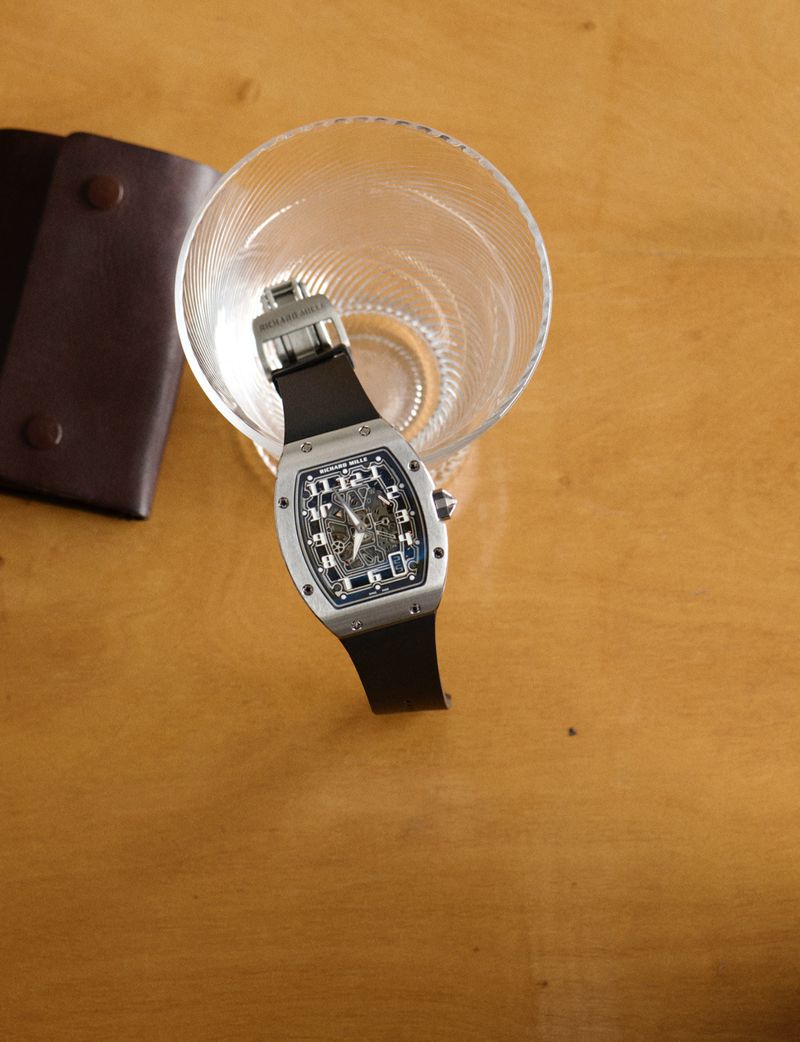Luxury silver wristwatch with a black band is displayed alongside a wallet and glass on a wood table in a detailed still life.