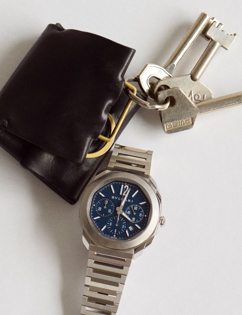 Luxury still life: silver Bvlgari watch with blue face, keys, and black leather wallet on a white surface, captured in a studio setting.