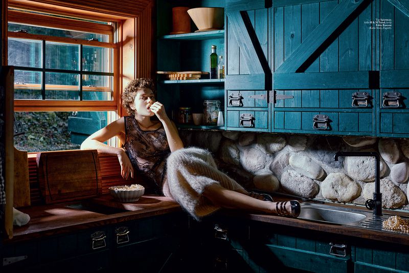 Woman in vintage dress poses in teal kitchen, eating from bowl of popcorn by window; stone backsplash and wooden details visible.