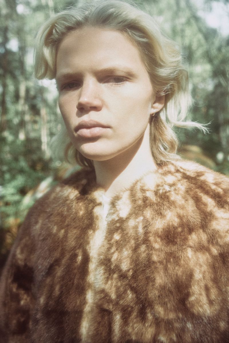 A fair-skinned adult with short blonde hair, wearing a fur coat stands in front of a blurred forest backdrop in natural light.