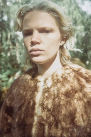 A fair-skinned adult with short blonde hair, wearing a fur coat stands in front of a blurred forest backdrop in natural light.