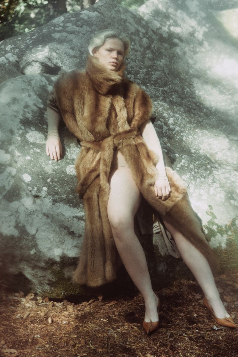 An adult woman leans against a textured rock in a forest setting, wearing a brown fur coat and tan heels in soft natural lighting.