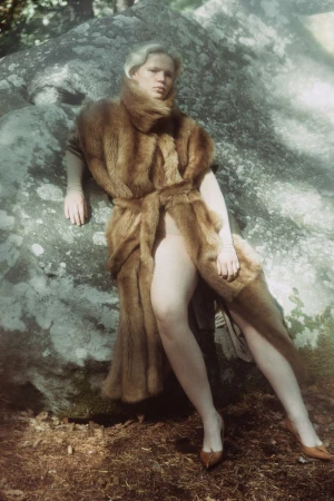 An adult woman leans against a textured rock in a forest setting, wearing a brown fur coat and tan heels in soft natural lighting.