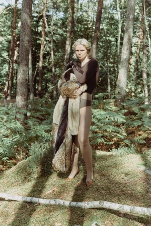 Blonde woman in nature wearing a brown bodysuit and fur coat standing on a grassy patch surrounded by green trees and ferns.