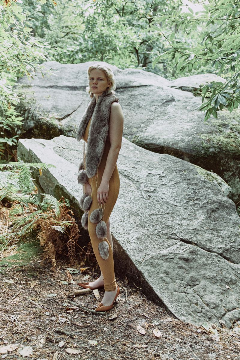 A woman in a fur stole and tan jumpsuit poses in a wooded area in front of a large rock, wearing pointed, tan colored flats.