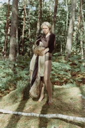 Model standing in a sunlit forest holding a textured fur wrap wearing a dark top and underwear
