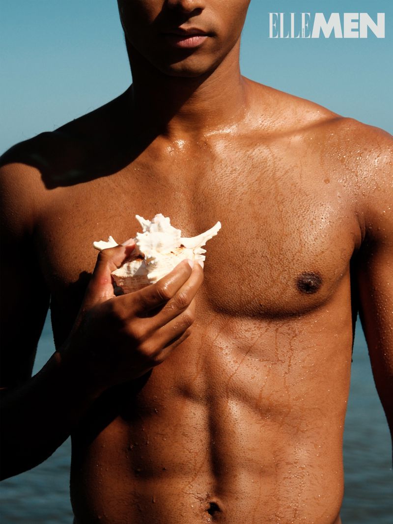 Dark-skinned shirtless man holds a seashell against a light blue background near water. The image includes 'ELLE MEN' text.