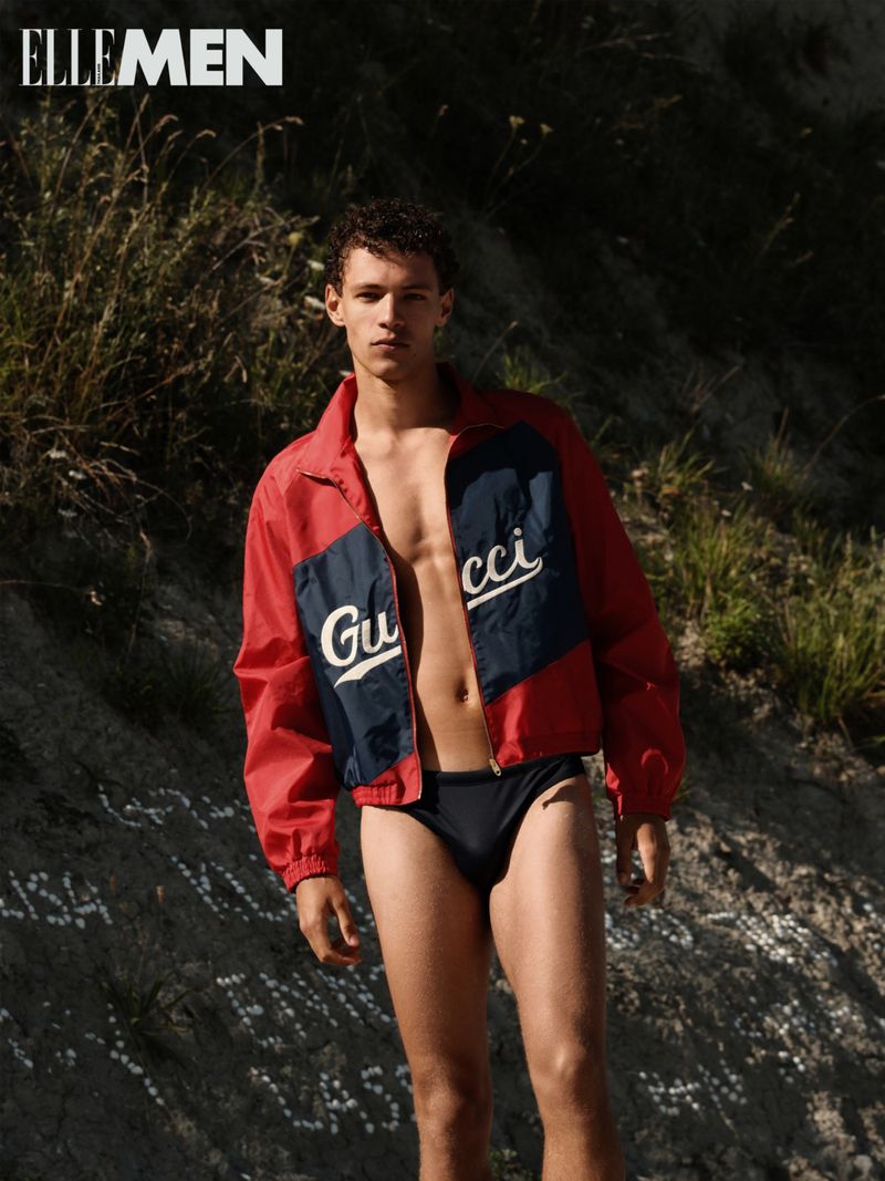 A young man in a Gucci jacket and black swim briefs poses outdoors with grassy terrain, showcased in a fashion editorial shot.