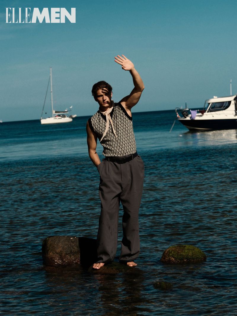 Man standing barefoot in water near boats. He wears a patterned vest, gray pants and a neck scarf while shielding his eyes on a sunny day.