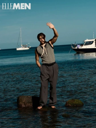 Man standing barefoot in water near boats. He wears a patterned vest, gray pants and a neck scarf while shielding his eyes on a sunny day.