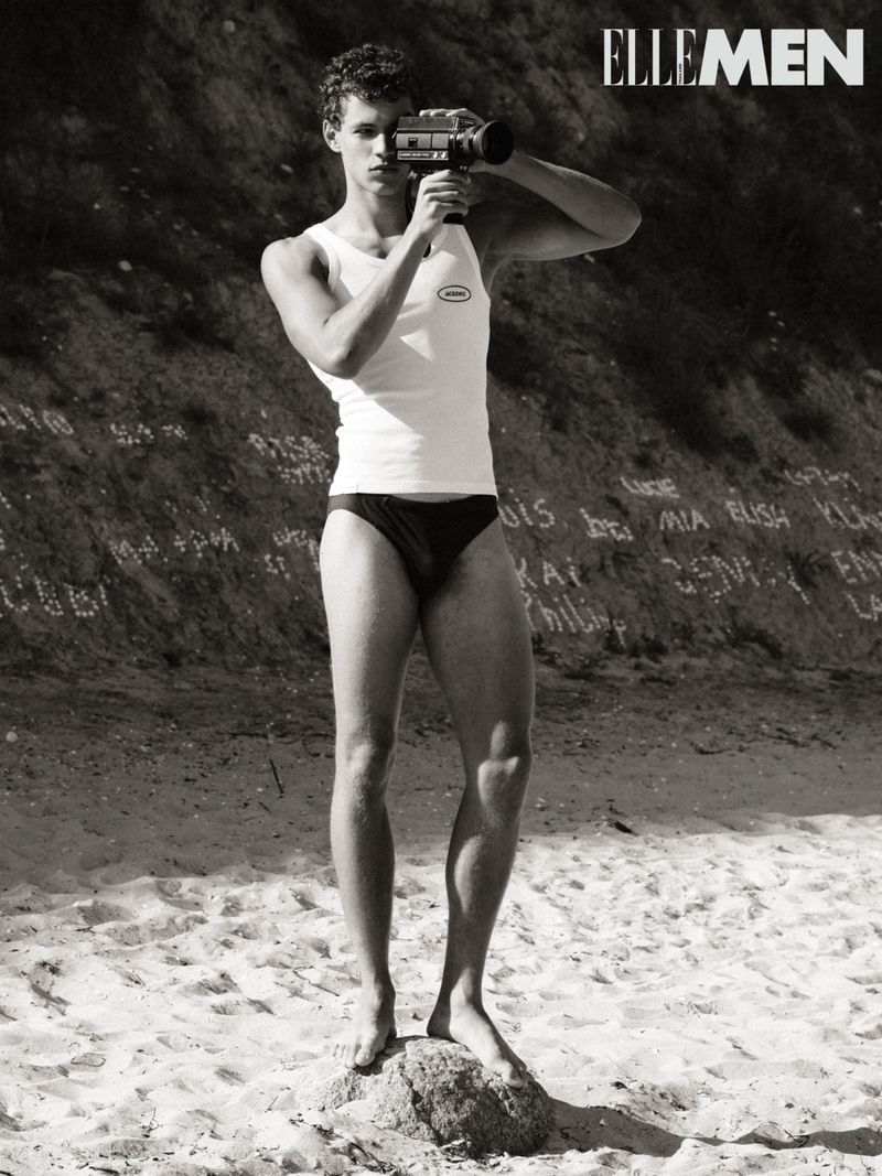 A young adult man in swimwear stands on a rock on a sandy beach, holding a vintage video camera. Black and white, seaside setting.