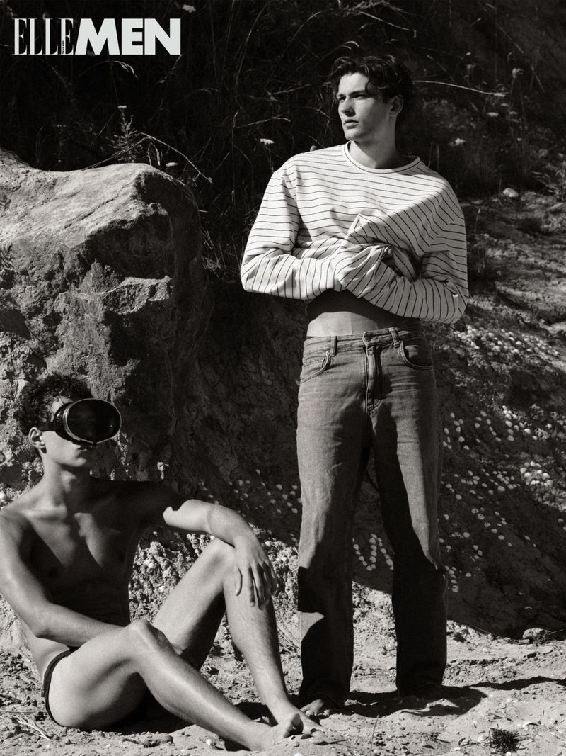 Black and white outdoor fashion shot of two adult men: one standing in jeans and a striped shirt, the other seated wearing swim goggles.