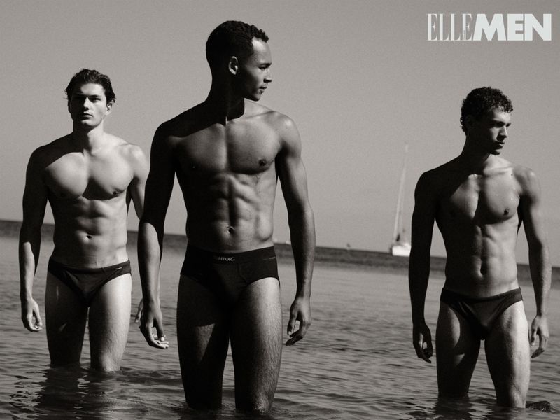 Three male fashion models pose in the ocean surf wearing swim briefs. The black-and-white image features a sailboat in the background.