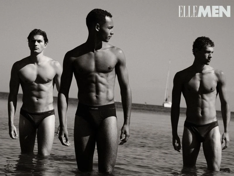 Three male fashion models pose in the ocean surf wearing swim briefs. The black-and-white image features a sailboat in the background.