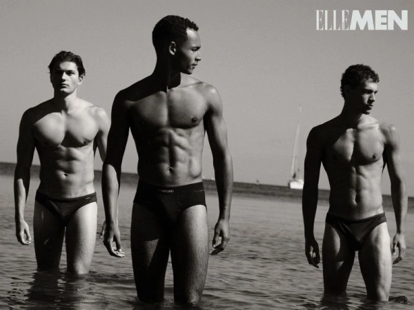 Three male fashion models pose in the ocean surf wearing swim briefs. The black-and-white image features a sailboat in the background.