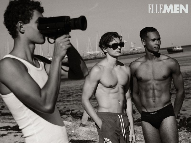 Black-and-white shot of three male models on a beach, one with a vintage camera, in an editorial fashion layout for Elle Men magazine.