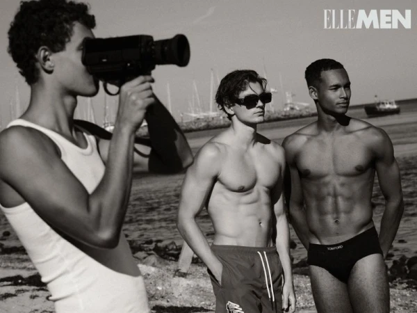 Black-and-white shot of three male models on a beach, one with a vintage camera, in an editorial fashion layout for Elle Men magazine.